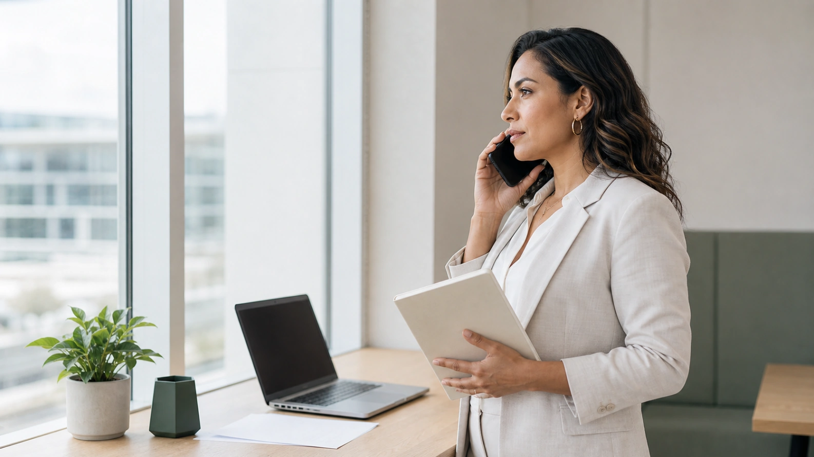 A person standing by a window on a phone call in warm light, a notepad in one hand, composed and thinking rather than rushing to answer